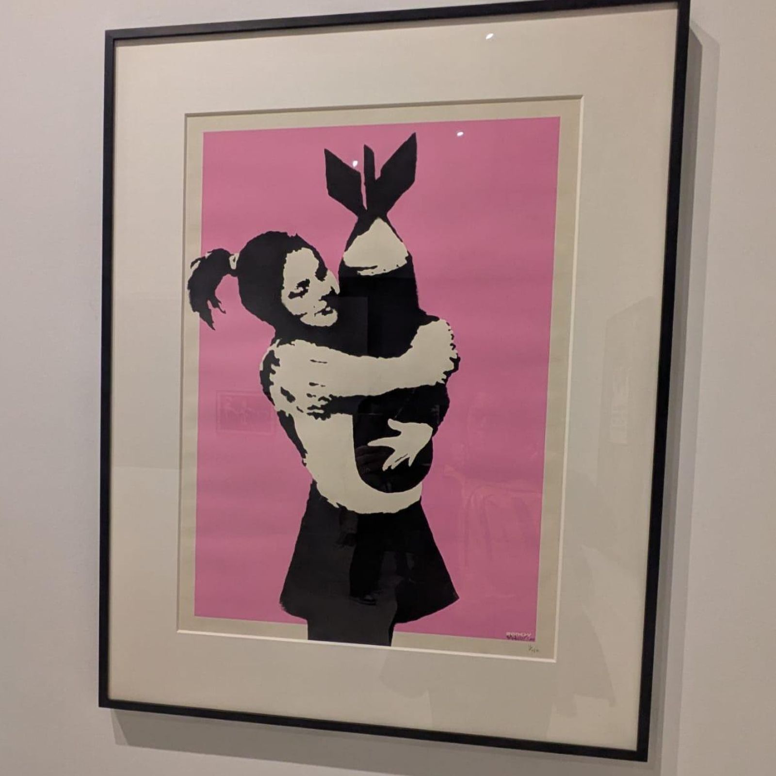 La imagen muestra la obra titulada "Bomb Hugger" o "Bomb Love" del artista urbano Banksy, que representa a una niña abrazando una bomba contra un fondo rosa.