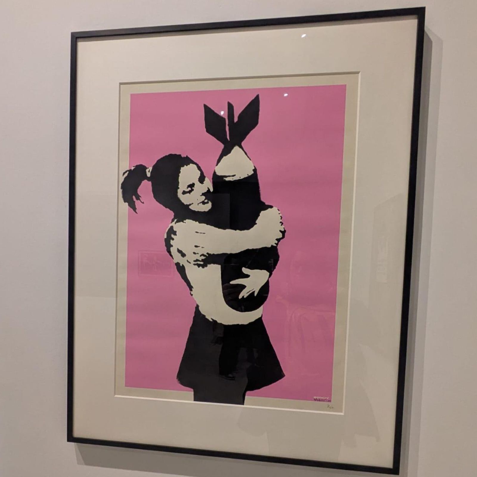 La imagen muestra la obra titulada "Bomb Hugger" o "Bomb Love" del artista urbano Banksy, que representa a una niña abrazando una bomba contra un fondo rosa.