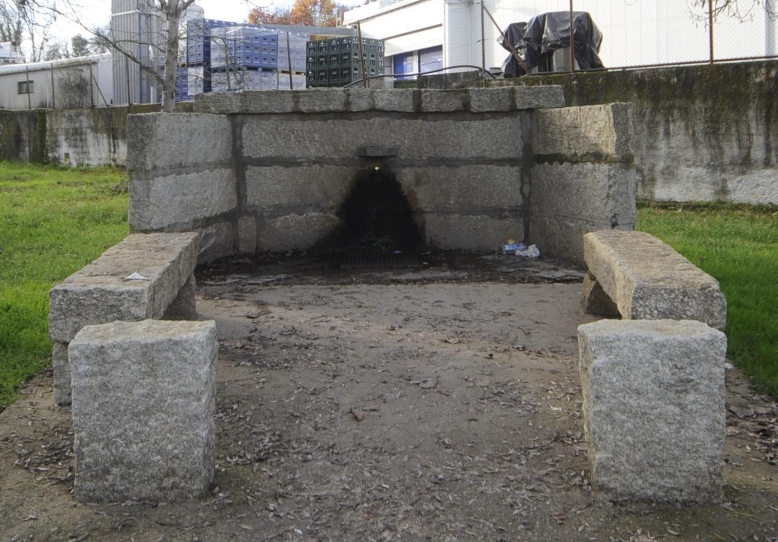 La actual fuente pública, muy tosca, situada en la parte posterior del manan- tial. Detrás, el muro de cierre de la fábrica y la nave, que rompe con el entorno.
