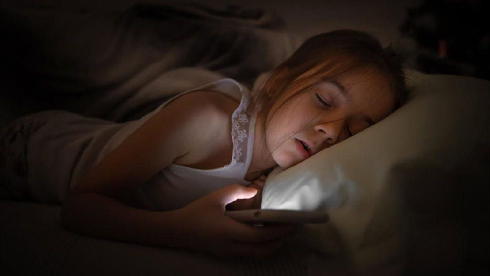 Una niña dormida con el móvil en la mano con la pantalla encendida.