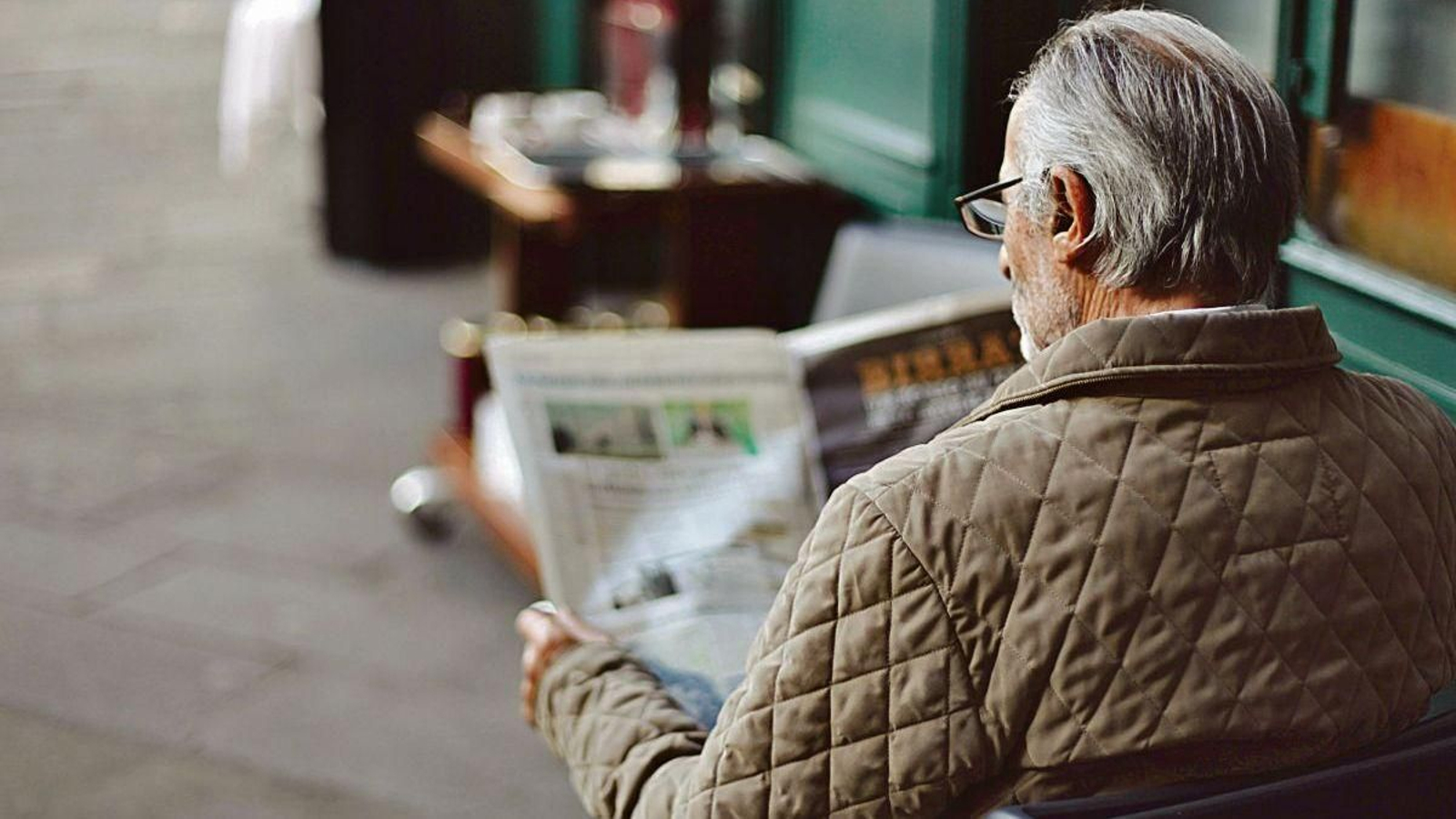 Un hombre sentado en una terraza en la calle lee un periódico.