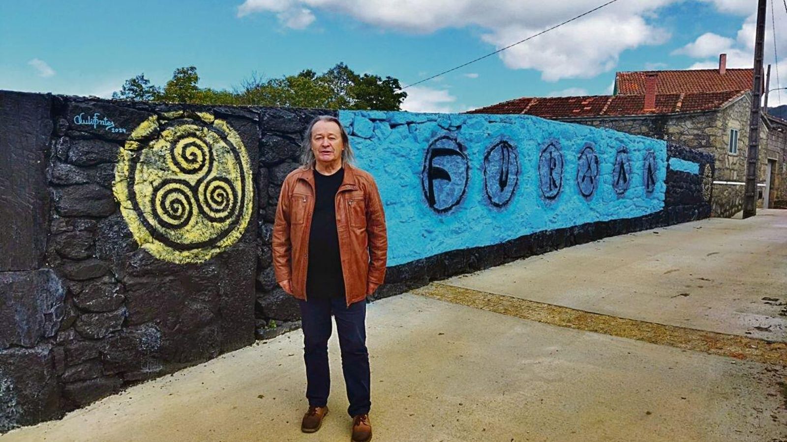 El pintor Xulio Fontes, frente al mural con el nombre del pueblo.