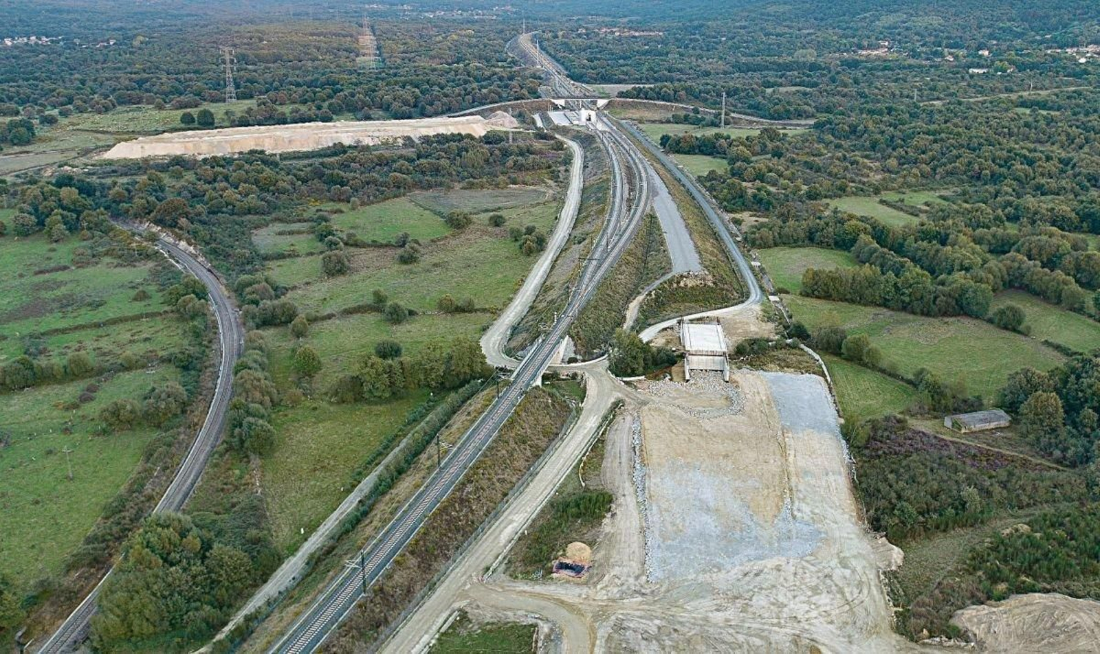 La variante exterior da continuidad a la línea de alta velocidad, pasado el cambiador de anchos, que vemos al fondo. A la izquierda, uno de los vertederos de material extraído de desmontes y el túnel. En primer plano, el pequeño viaducto sobre el río Taboadela.
