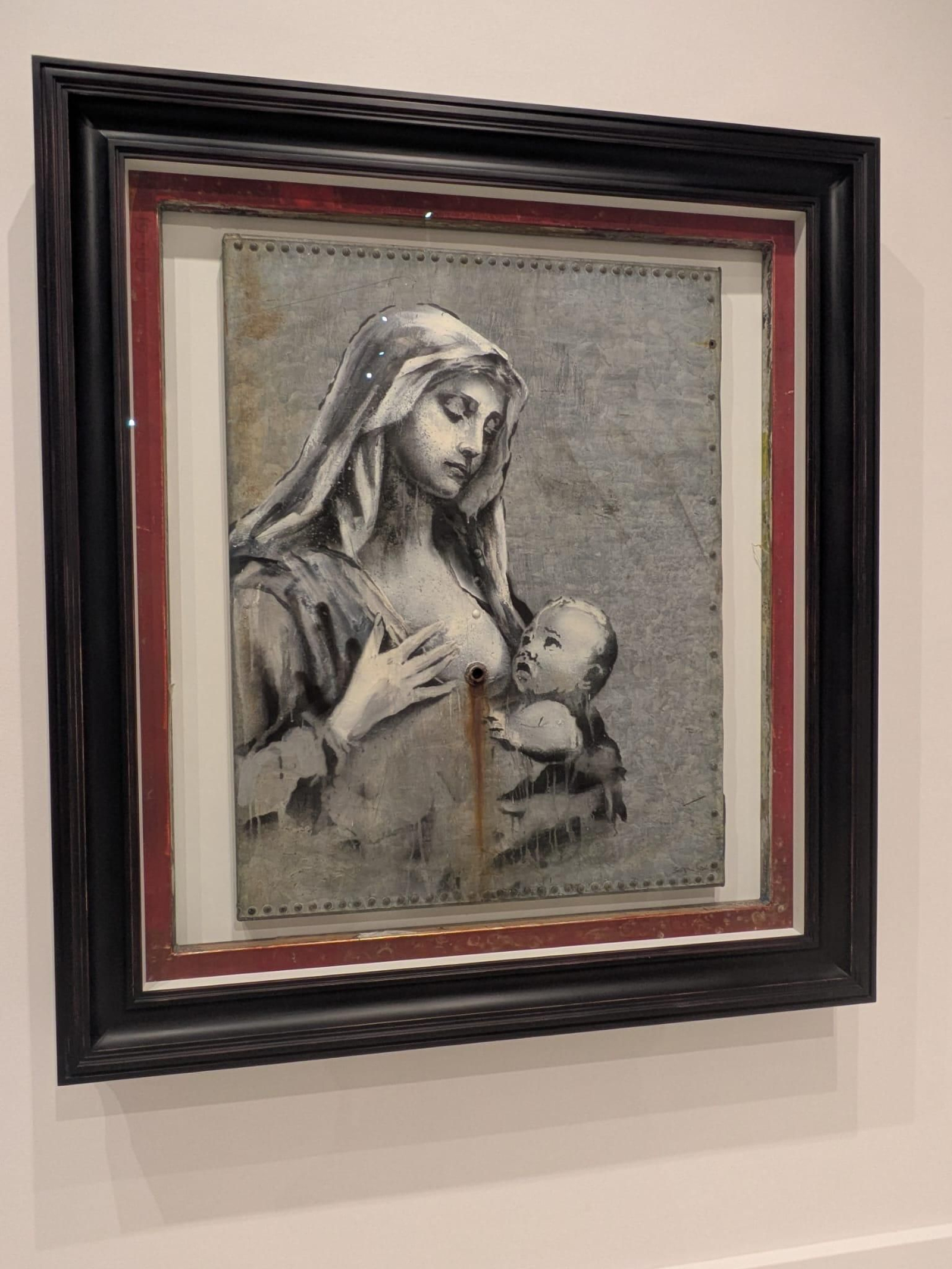 A lo largo de los años, Banksy ha creado diversas versiones del motivo de la Virgen María y el Niño Jesús. Esta versión sobre una placa de acero galvanizado es una alegoría a que ni en las guerras más crudas falta lo básico que una madre ofrece a su hijo.  En esta impactante "Madonna and Child" , una Virgen María amamanta al niño Jesús, obra pintada sobre chatarra y donde el pezón es un tubo oxidado.