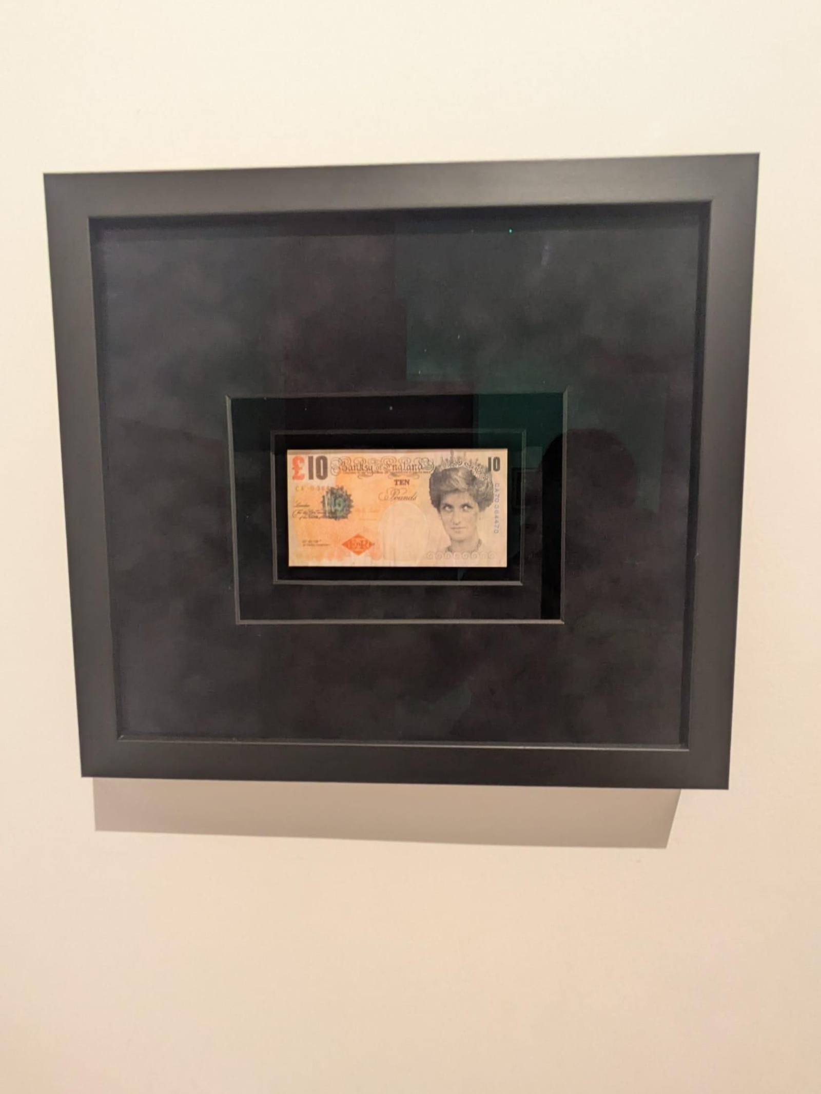 Esta imagen muestra una obra de arte enmarcada titulada "Di-Faced Tenner" creada por Banksy, que consiste en un billete falso de 10 libras esterlinas con el rostro de la princesa Diana en lugar de la reina Isabel II.